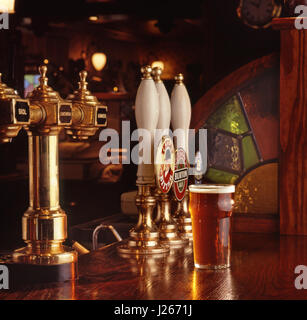 Pint of bitter served on the wooden bar surface of a warm, typical, traditional inviting English public house, with beer and lager pump handles behind Stock Photo