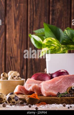 Vertical photo of three slices of raw pork meat which are placed on dark wooden board with whole pepper, milled red capsicum, few pieces of quail eggs Stock Photo