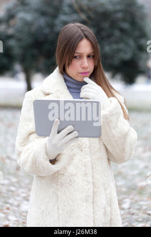 Young woman in a fur coat, standing in the park and looking thoughtfully into tablets Stock Photo