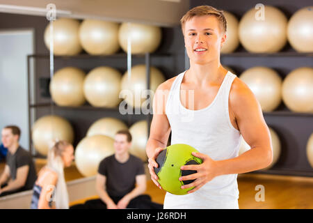Smiling Man Holding Medicine Ball While Friends Resting In Gym Stock Photo