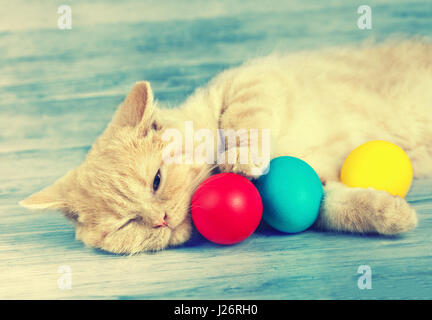 Little ginger kitten sleeping on blue grunge wooden table and holding three colored eggs. Easter concept scene. Retro color Stock Photo