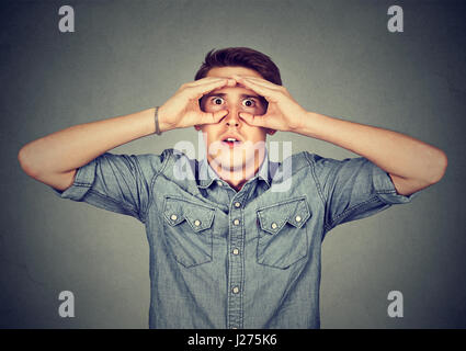 Closeup portrait young stunned curious man, peeking looking through fingers like binoculars searching something future forecast isolated grey backgrou Stock Photo