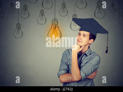 Thoughtful graduate student young man in cap gown looking up at bright light bulb thinking isolated gray wall background. Graduation ceremony future c Stock Photo