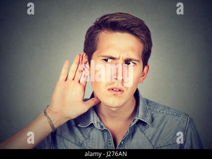 Closeup portrait hard of hearing man placing hand on ear asking someone to speak up or listening carefully to a gossip isolated on gray wall backgroun Stock Photo