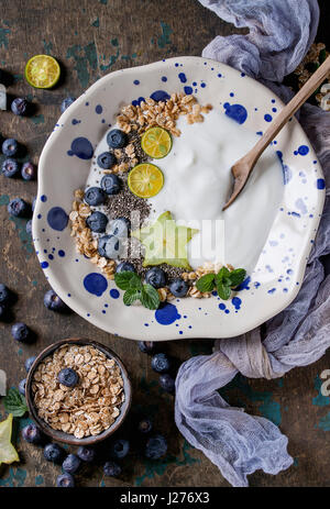Smoothie bowl healthy breakfast. Yogurt with blueberries, granola, chia seeds, lime, mint and carambola on textile gauze over dark wooden texture back Stock Photo
