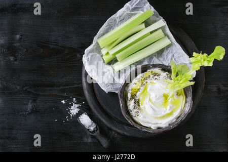Fresh celery on paper with yogurt and olive oil dip in ceramic bowl, served with sea salt on wood round chopping board over black wooden burnt backgro Stock Photo