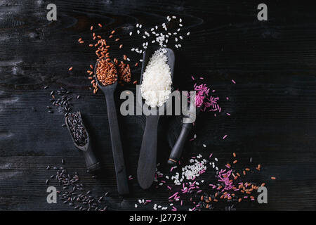 Variety assortment of raw uncooked colorful rice white, black, brown, pink in black spoons and scoop over burnt wooden background. Top view with space Stock Photo