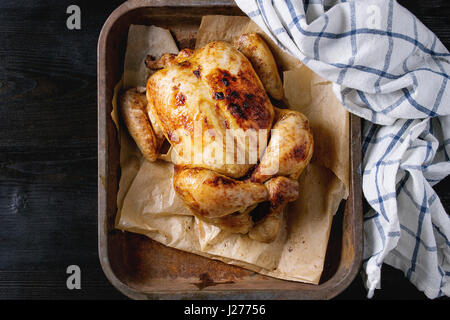 Grilled baked whole organic chicken on backing paper in old oven tray with white kitchen towel over black burnt wooden background. Top view with space Stock Photo