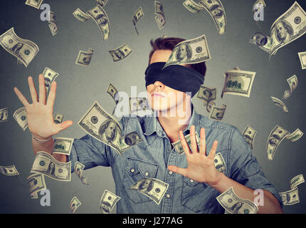 Blindfolded young businessman trying to catch dollar bills banknotes flying in the air isolated on gray wall background. Financial corporate success o Stock Photo