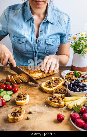 A woman slicing French baguette photographed from front view. Olive tapenade on bread slices, olives, pickled baby corns, strawberries, tomatoes, cucu Stock Photo