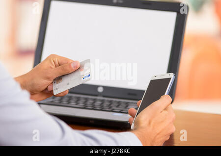 Closeup of young mans hands holding smartphone up, Visa card in other hand with laptop computer sitting on desk Stock Photo