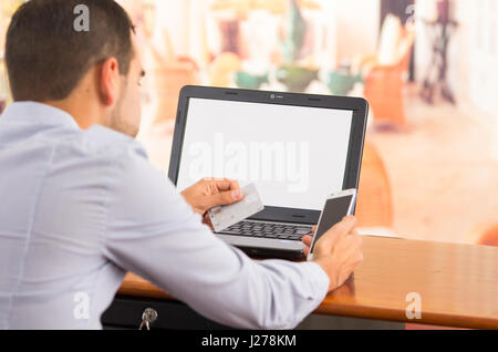 Closeup of young mans hands holding smartphone up, Visa card in other hand with laptop computer sitting on desk Stock Photo