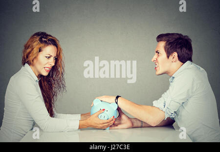 Finances in divorce concept. Wife and husband can not make settlement holding piggy bank sitting at table looking at each other with hatred Stock Photo