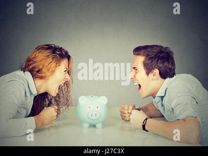 Finances in divorce concept. Wife and husband can not make settlement screaming piggy bank in-between sitting at table looking at each other with hatr Stock Photo