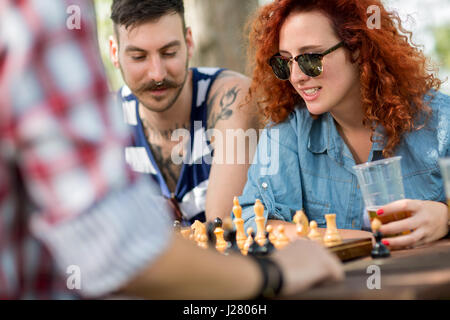 Curly ginger haired female plays chess with male friends at camp in nature Stock Photo