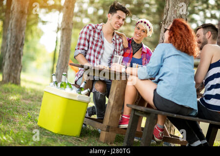 Girls and guys sits on bench at wooden table in forest, drink beer and make jokes Stock Photo