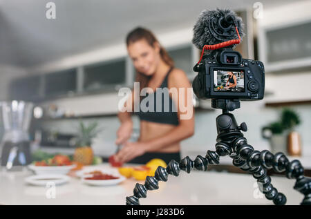 Young lady in sports wear recording video on tripod mounted camera in kitchen. Woman cutting fruits on kitchen table. Stock Photo