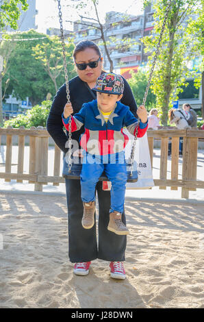 A mother pushes he young son who is playing on a swing in a children's playground. Stock Photo