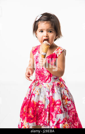 indian adorable infant or girl child licking or eating chocolate ice cream in cone cone and showing happiness, isolated over white background Stock Photo