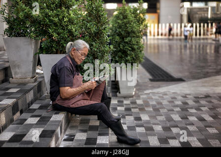Woman taking a break from work and sat reading the newspaper outside on the street.  Bangkok, Thailand, Stock Photo