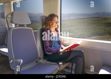 red hair smiling woman dressed in purple and blue, traveling by train sitting reading red open book Stock Photo