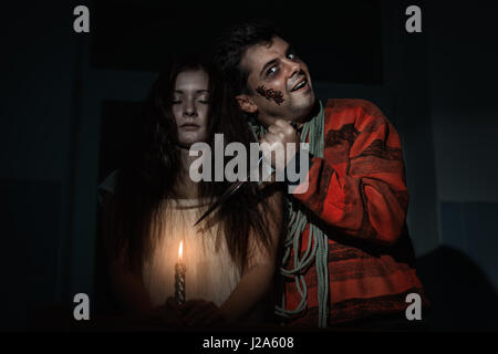 Creepy man and woman are standing in the room, he had a knife and weighs a rope around his neck. Stock Photo