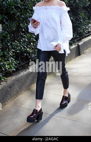 LONDON - FEBRUARY, 2017: Low section of woman wearing white off the shoulder blouse walking in street using phone during London Fashion Week Stock Photo