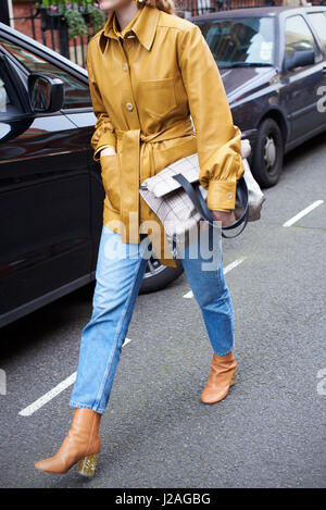 LONDON - FEBRUARY, 2017: Low section of woman wearing jeans and yellow leather jacket, holding large handbag in street during London Fashion Week, vertical Stock Photo