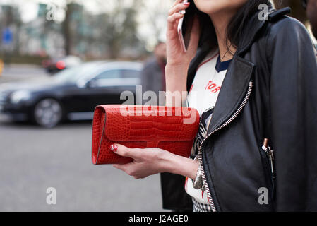 LONDON - FEBRUARY, 2017: Mid section close up of woman wearing a black leather bike jacket, holding a red Hermes purse and using smartphone in the street during London Fashion Week, horizontal Stock Photo