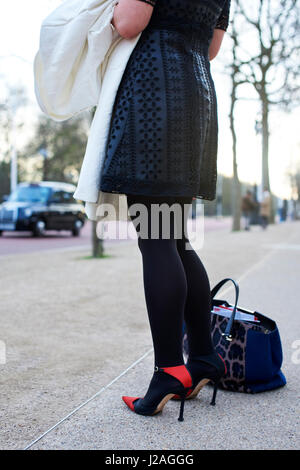 LONDON - FEBRUARY, 2017: Low section of woman wearing black and red stiletto heel shoes and black patterned dress standing at roadside during London Fashion Week, vertical, back view Stock Photo