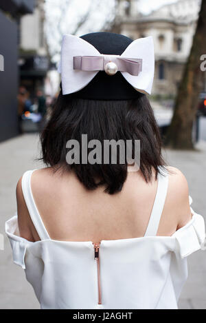 LONDON - FEBRUARY, 2017: Back view of woman wearing brimless hat with large bow decoration and white dress standing in the street during London Fashion Week, vertical Stock Photo