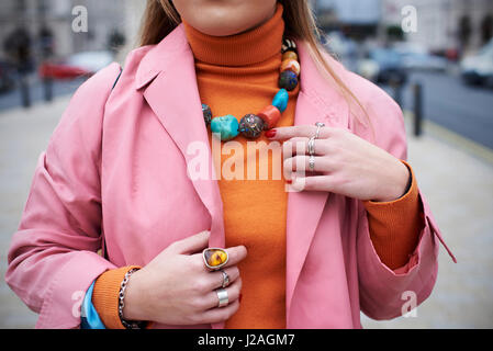 LONDON - FEBRUARY, 2017: Mid section of woman wearing pink coat and orange sweater with chunky stone necklace and rings standing in a street during London Fashion Week, horizontal Stock Photo