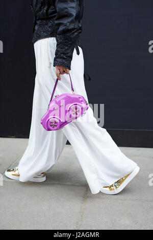 LONDON - FEBRUARY, 2017: Mid section of woman with blue hair wearing leather jacket and baggy white trousers holding purple car-shaped handbag walking in a street during London Fashion Week, vertical, side view Stock Photo