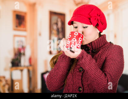 Pretty young brunette woman wearing red sweater and beenie, taking a sip from cup of hot beverage Stock Photo
