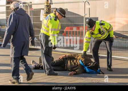 Two Metropolitan Police officers assisting an inebriated man who is asleep on the pavement on a sunny day.
