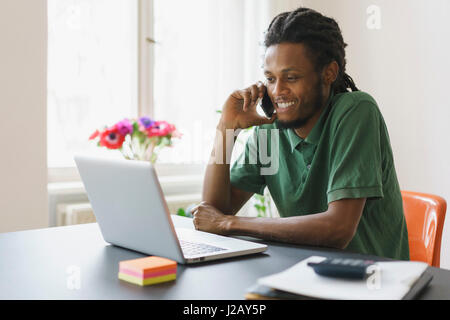 Happy man looking at laptop while using smart phone in house Stock Photo