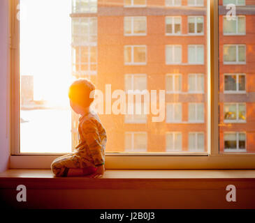 Side view of boy looking out while kneeling on window sill at home Stock Photo