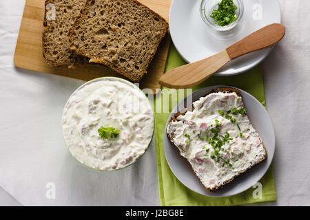 Slice of wholegrain rye bread with delicate light and fresh cottage cheese spread sprinkled with chives and parsley. Wooden spreading knife and some e Stock Photo