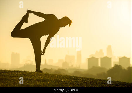 Silhouette of a slim man stretching in front of a misty golden sunrise view of the city skyline from the grass of a park hill Stock Photo