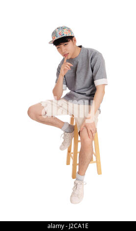 A young Korean teenager sitting on a chair in shorts and a cap with his finger on his chin, isolated for white background. Stock Photo