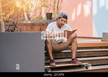 Young Asian man relaxing on stairs outside reading a book Stock Photo