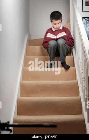 London,Uk-schoolboy in school uniform reads book sitting on stairs at home Stock Photo