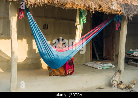 A woman, her baby sleeping in a hammock, is sitting in front of a typical farm house in the village Pandavnagar in Chitwan National Park Stock Photo