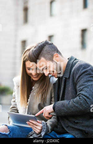 Portrait of beautiful smilling young couple using a digital tablet and enjoying together, sitting on a bench. Modern relationship concept. Websurfing. Stock Photo