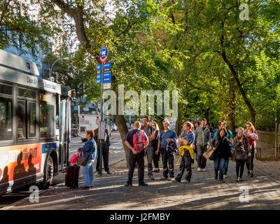 A diversified crowd of travelers waits at a southbound bus stop by Central Park on Fifth Avenue, NY City, on a sunny summer afternoon as a bus arrives. Note sign listing different buses. Stock Photo