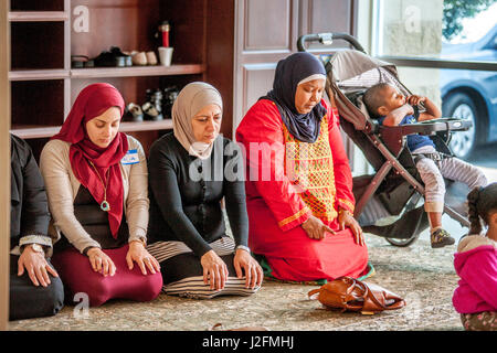 Wearing a hijabs or head scarves in a separate room, Muslim women kneel in Abrahamic prayer while listening to the sermon or Khutbah during religious services at an Anaheim, CA, mosque. Note traditional Middle Eastern clothing and child in stroller. Stock Photo