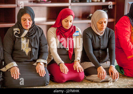Wearing a hijabs or head scarves in a separate room, Muslim women kneel in Abrahamic prayer while listening to the sermon or Khutbah during religious services at an Anaheim, CA, mosque. Note traditional Middle Eastern clothing. Stock Photo
