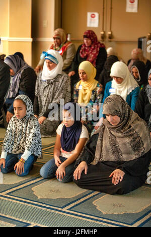 Wearing a hijabs or head scarves in a separate area, Muslim women and girls kneel in Abrahamic prayer while listening to the sermon or Khutbah during religious services at an Anaheim, CA, mosque. Note traditional Middle Eastern clothing. Stock Photo