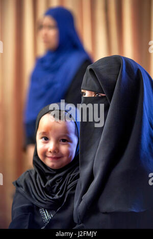 A Muslim mother wearing a face-covering burqua holds her toddler son in traditional Middle Eastern clothing at an Anaheim, CA, mosque. Note woman in background wearing a hijab, the traditional Muslim female head scarf. Stock Photo