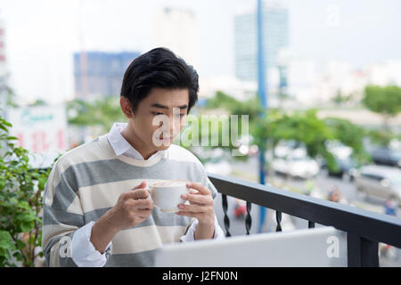 Handsome asian young man working on laptop and smiling while enjoying coffee in cafe Stock Photo